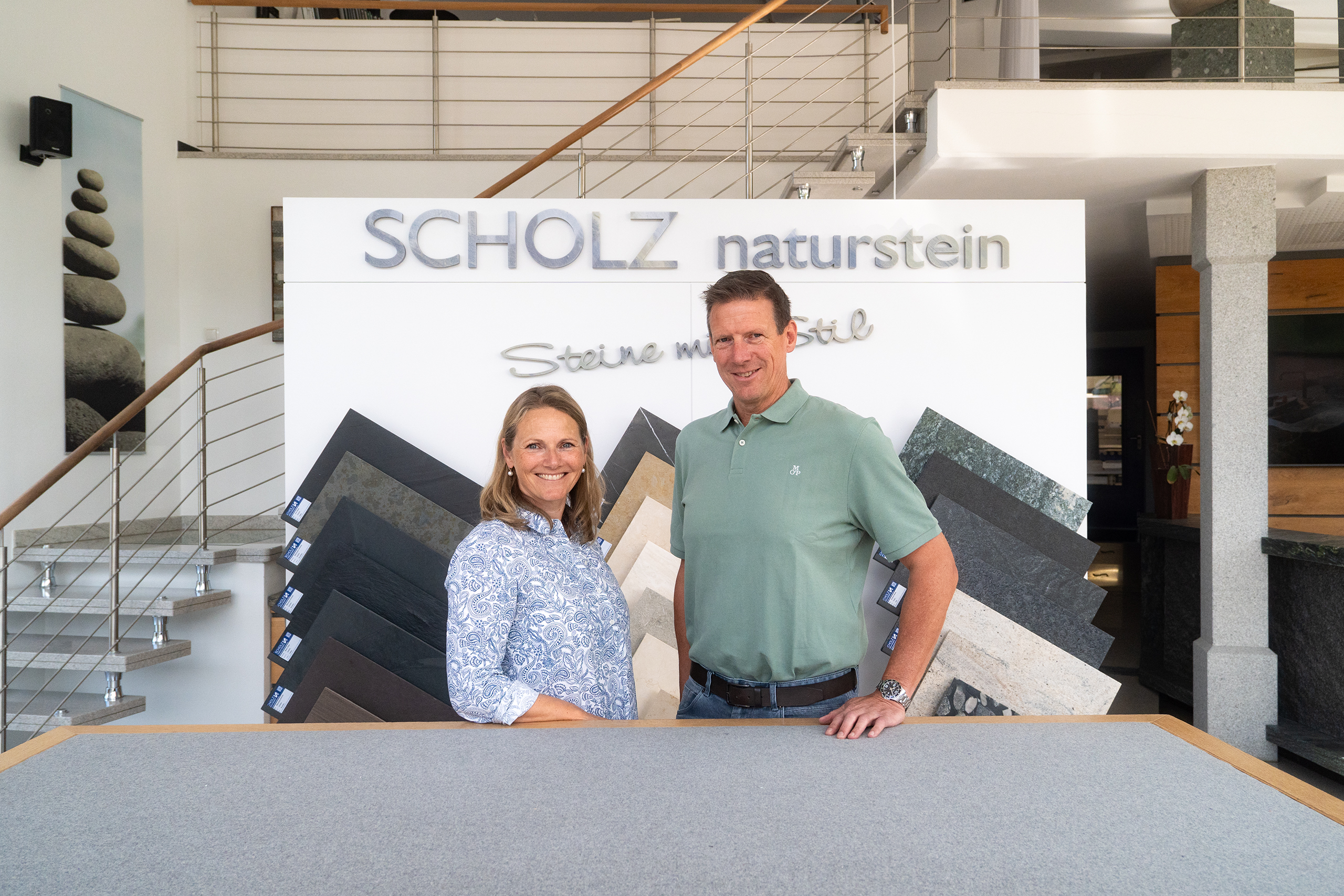 Tanja und Andreas Scholz stehen lächelnd in der Ausstellung vor der Logowand „Scholz Naturstein – Steine mit Stil" und Natursteinmustern