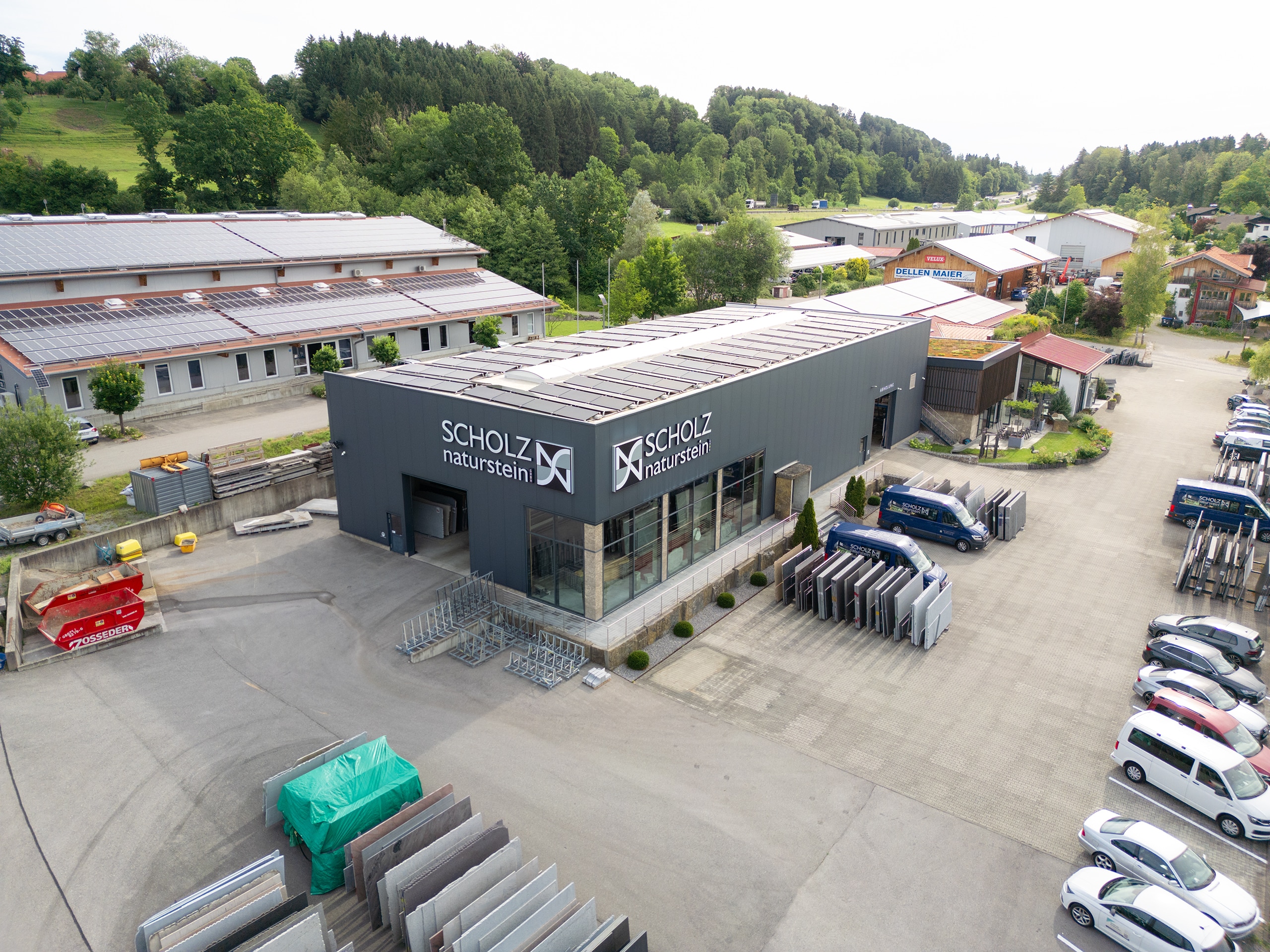 Luftaufnahme der Scholz-Naturstein-Halle mit Schriftzug, davor Parkplatz, Firmentransporter und gelagerte Natursteinplatten